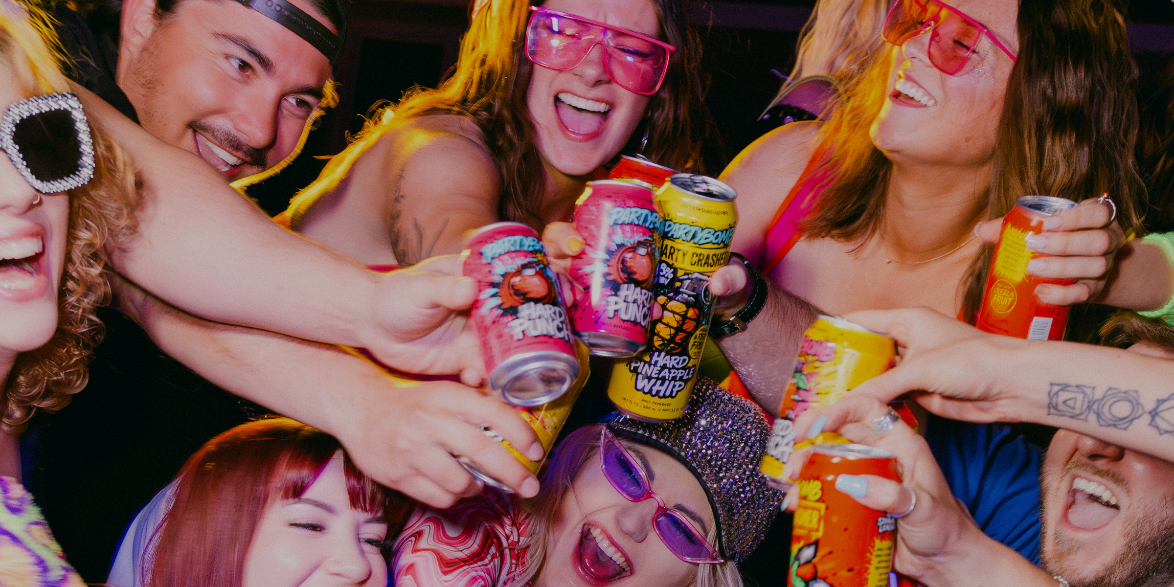 A group of smiling, laughing people in a dimly lit environment reaching toward colorful beverage cans. Several people are wearing pink sunglasses, and the scene is illuminated by warm, low-light ambient lighting.